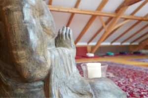 Meditationsraum mit Orientteppich und Dachschrägen. Im Vordergrund ein Ausschnitt einer Buddha-Statue.