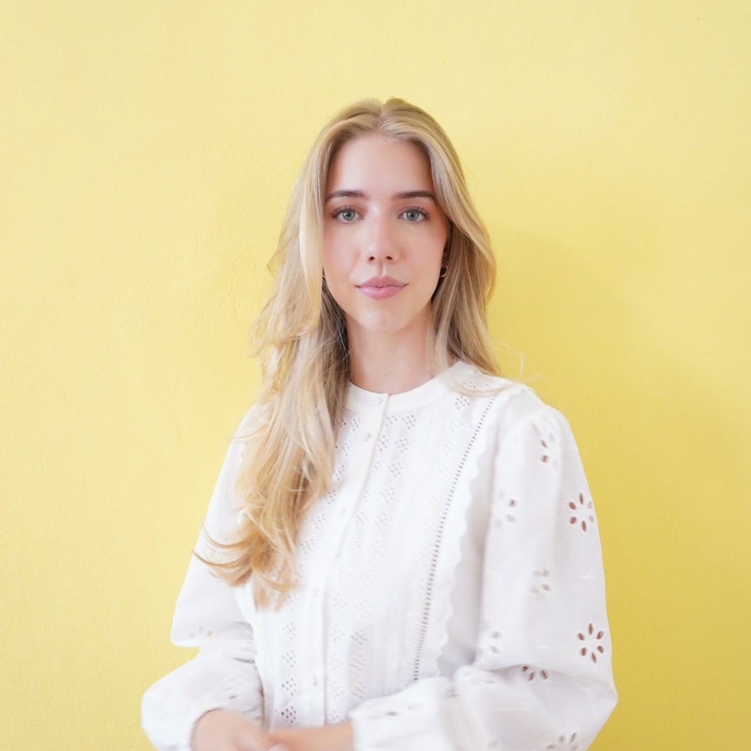 Alisia steht vor einer gelben Wand, trägt eine weiße Bluse und lächelt in die Kamera